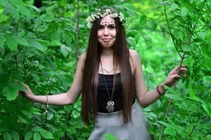 Portrait of an emotional young girl with a floral wreath on her head and shiny ornaments on her forehead. Cute brunette posing in a burgeoning beautiful forest in the daytime on a fine day photo