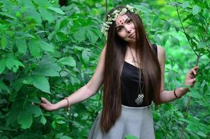 Portrait of an emotional young girl with a floral wreath on her head and shiny ornaments on her forehead. Cute brunette posing in a burgeoning beautiful forest in the daytime on a fine day photo