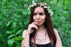 una foto del bosque de una hermosa joven morena de apariencia europea con ojos marrones oscuros y labios grandes. en la cabeza de la niña lleva una corona de flores, en la frente adornos brillantes