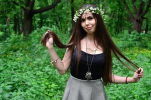 una foto del bosque de una hermosa joven morena de apariencia europea con ojos marrones oscuros y labios grandes. en la cabeza de la niña lleva una corona de flores, en la frente adornos brillantes