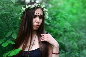 retrato de una joven emocional con una corona floral en la cabeza y adornos brillantes en la frente. linda morena posando en un hermoso bosque floreciente durante el día en un buen día foto