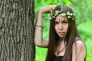 A forest picture of a beautiful young brunette of European appearance with dark brown eyes and large lips. On the girl's head is wearing a floral wreath, on her forehead shiny decorations photo