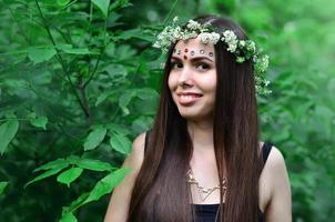 Portrait of an emotional young girl with a floral wreath on her head and shiny ornaments on her forehead. Cute brunette posing in a burgeoning beautiful forest in the daytime on a fine day photo