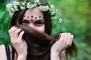 una foto del bosque de una hermosa joven morena de apariencia europea con ojos marrones oscuros y labios grandes. en la cabeza de la niña lleva una corona de flores, en la frente adornos brillantes