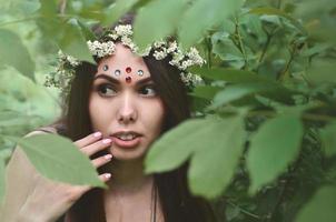 una foto del bosque de una hermosa joven morena de apariencia europea con ojos marrones oscuros y labios grandes. en la cabeza de la niña lleva una corona de flores, en la frente adornos brillantes