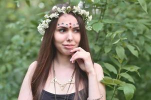 una foto del bosque de una hermosa joven morena de apariencia europea con ojos marrones oscuros y labios grandes. en la cabeza de la niña lleva una corona de flores, en la frente adornos brillantes