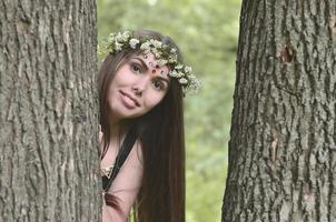 Portrait of an emotional young girl with a floral wreath on her head and shiny ornaments on her forehead. Cute brunette posing in a burgeoning beautiful forest in the daytime on a fine day photo