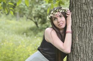 retrato de una joven emocional con una corona floral en la cabeza y adornos brillantes en la frente. linda morena posando en un hermoso bosque floreciente durante el día en un buen día foto