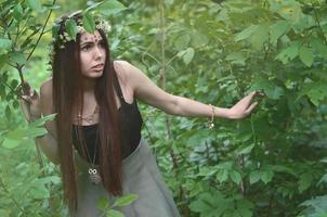 una foto del bosque de una hermosa joven morena de apariencia europea con ojos marrones oscuros y labios grandes. en la cabeza de la niña lleva una corona de flores, en la frente adornos brillantes