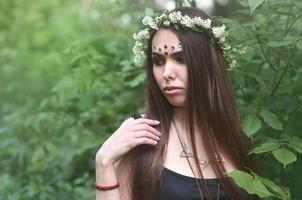 Portrait of an emotional young girl with a floral wreath on her head and shiny ornaments on her forehead. Cute brunette posing in a burgeoning beautiful forest in the daytime on a fine day photo