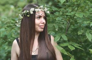 una foto del bosque de una hermosa joven morena de apariencia europea con ojos marrones oscuros y labios grandes. en la cabeza de la niña lleva una corona de flores, en la frente adornos brillantes