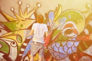 Photo in the process of drawing a graffiti pattern on an old concrete wall. Young long-haired blond guy draws an abstract drawing of different colors. Street art and vandalism concept