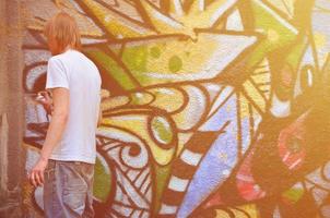 foto en el proceso de dibujar un patrón de graffiti en un antiguo muro de hormigón. un joven rubio de pelo largo dibuja un dibujo abstracto de diferentes colores. concepto de arte callejero y vandalismo