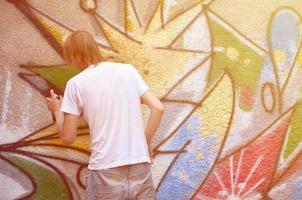 Photo in the process of drawing a graffiti pattern on an old concrete wall. Young long-haired blond guy draws an abstract drawing of different colors. Street art and vandalism concept