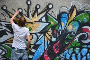 Photo in the process of drawing a graffiti pattern on an old concrete wall. Young long-haired blond guy draws an abstract drawing of different colors. Street art and vandalism concept