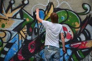 foto en el proceso de dibujar un patrón de graffiti en un antiguo muro de hormigón. un joven rubio de pelo largo dibuja un dibujo abstracto de diferentes colores. concepto de arte callejero y vandalismo