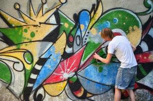 Photo in the process of drawing a graffiti pattern on an old concrete wall. Young long-haired blond guy draws an abstract drawing of different colors. Street art and vandalism concept