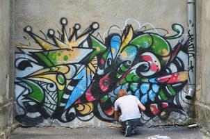 foto en el proceso de dibujar un patrón de graffiti en un antiguo muro de hormigón. un joven rubio de pelo largo dibuja un dibujo abstracto de diferentes colores. concepto de arte callejero y vandalismo