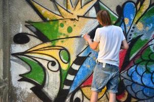 Photo of a young guy in denim shorts and a white shirt. The guy draws on the graffiti wall a drawing with aerosol paints of various colors. The concept of hooliganism and damage to property