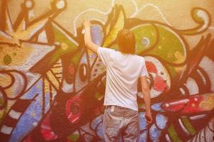 foto en el proceso de dibujar un patrón de graffiti en un antiguo muro de hormigón. un joven rubio de pelo largo dibuja un dibujo abstracto de diferentes colores. concepto de arte callejero y vandalismo