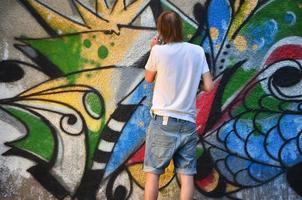 foto en el proceso de dibujar un patrón de graffiti en un antiguo muro de hormigón. un joven rubio de pelo largo dibuja un dibujo abstracto de diferentes colores. concepto de arte callejero y vandalismo