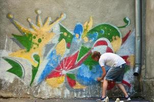 Photo of a young guy in denim shorts and a white shirt. The guy draws on the graffiti wall a drawing with aerosol paints of various colors. The concept of hooliganism and damage to property