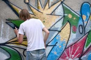 foto en el proceso de dibujar un patrón de graffiti en un antiguo muro de hormigón. un joven rubio de pelo largo dibuja un dibujo abstracto de diferentes colores. concepto de arte callejero y vandalismo