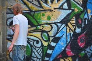 foto en el proceso de dibujar un patrón de graffiti en un antiguo muro de hormigón. un joven rubio de pelo largo dibuja un dibujo abstracto de diferentes colores. concepto de arte callejero y vandalismo