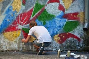 Photo in the process of drawing a graffiti pattern on an old concrete wall. Young long-haired blond guy draws an abstract drawing of different colors. Street art and vandalism concept