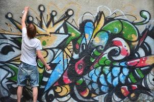 Photo in the process of drawing a graffiti pattern on an old concrete wall. Young long-haired blond guy draws an abstract drawing of different colors. Street art and vandalism concept