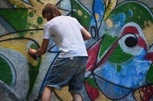 foto en el proceso de dibujar un patrón de graffiti en un antiguo muro de hormigón. un joven rubio de pelo largo dibuja un dibujo abstracto de diferentes colores. concepto de arte callejero y vandalismo