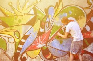 Photo in the process of drawing a graffiti pattern on an old concrete wall. Young long-haired blond guy draws an abstract drawing of different colors. Street art and vandalism concept