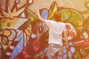 Photo of a young guy in denim shorts and a white shirt. The guy draws on the graffiti wall a drawing with aerosol paints of various colors. The concept of hooliganism and damage to property