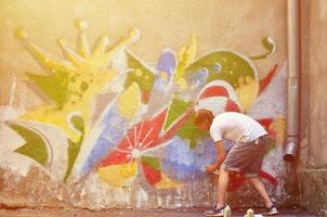 Photo of a young guy in denim shorts and a white shirt. The guy draws on the graffiti wall a drawing with aerosol paints of various colors. The concept of hooliganism and damage to property