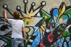 Photo of a young guy in denim shorts and a white shirt. The guy draws on the graffiti wall a drawing with aerosol paints of various colors. The concept of hooliganism and damage to property