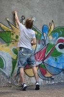 foto en el proceso de dibujar un patrón de graffiti en un antiguo muro de hormigón. un joven rubio de pelo largo dibuja un dibujo abstracto de diferentes colores. concepto de arte callejero y vandalismo