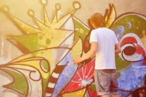 foto de un joven con pantalones cortos de mezclilla y una camisa blanca. el chico dibuja en la pared de graffiti un dibujo con pinturas en aerosol de varios colores. el concepto de vandalismo y daños a la propiedad