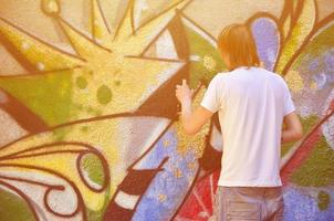 foto en el proceso de dibujar un patrón de graffiti en un antiguo muro de hormigón. un joven rubio de pelo largo dibuja un dibujo abstracto de diferentes colores. concepto de arte callejero y vandalismo