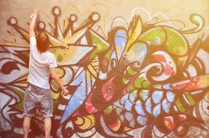 Photo in the process of drawing a graffiti pattern on an old concrete wall. Young long-haired blond guy draws an abstract drawing of different colors. Street art and vandalism concept