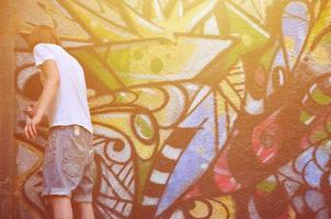 Photo in the process of drawing a graffiti pattern on an old concrete wall. Young long-haired blond guy draws an abstract drawing of different colors. Street art and vandalism concept