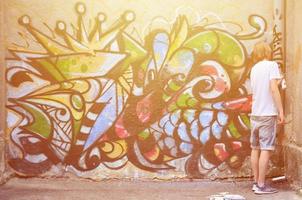 Photo in the process of drawing a graffiti pattern on an old concrete wall. Young long-haired blond guy draws an abstract drawing of different colors. Street art and vandalism concept