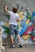 Photo in the process of drawing a graffiti pattern on an old concrete wall. Young long-haired blond guy draws an abstract drawing of different colors. Street art and vandalism concept