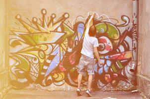 Photo of a young guy in denim shorts and a white shirt. The guy draws on the graffiti wall a drawing with aerosol paints of various colors. The concept of hooliganism and damage to property