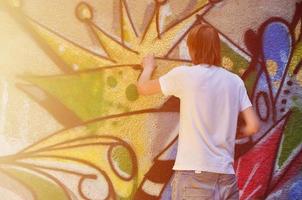 Photo of a young guy in denim shorts and a white shirt. The guy draws on the graffiti wall a drawing with aerosol paints of various colors. The concept of hooliganism and damage to property