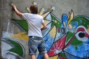 foto en el proceso de dibujar un patrón de graffiti en un antiguo muro de hormigón. un joven rubio de pelo largo dibuja un dibujo abstracto de diferentes colores. concepto de arte callejero y vandalismo