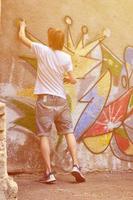 Photo in the process of drawing a graffiti pattern on an old concrete wall. Young long-haired blond guy draws an abstract drawing of different colors. Street art and vandalism concept