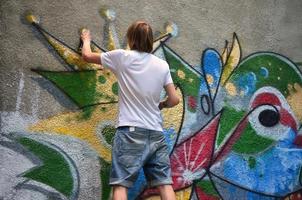 foto de un joven con pantalones cortos de mezclilla y una camisa blanca. el chico dibuja en la pared de graffiti un dibujo con pinturas en aerosol de varios colores. el concepto de vandalismo y daños a la propiedad