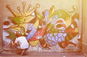 foto en el proceso de dibujar un patrón de graffiti en un antiguo muro de hormigón. un joven rubio de pelo largo dibuja un dibujo abstracto de diferentes colores. concepto de arte callejero y vandalismo