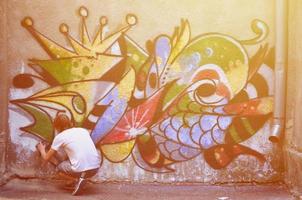 Photo in the process of drawing a graffiti pattern on an old concrete wall. Young long-haired blond guy draws an abstract drawing of different colors. Street art and vandalism concept