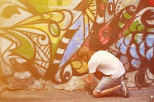 Photo in the process of drawing a graffiti pattern on an old concrete wall. Young long-haired blond guy draws an abstract drawing of different colors. Street art and vandalism concept